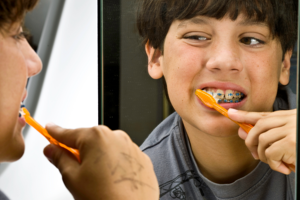 small kid using an orange toothbrush to clean his teeth and braces